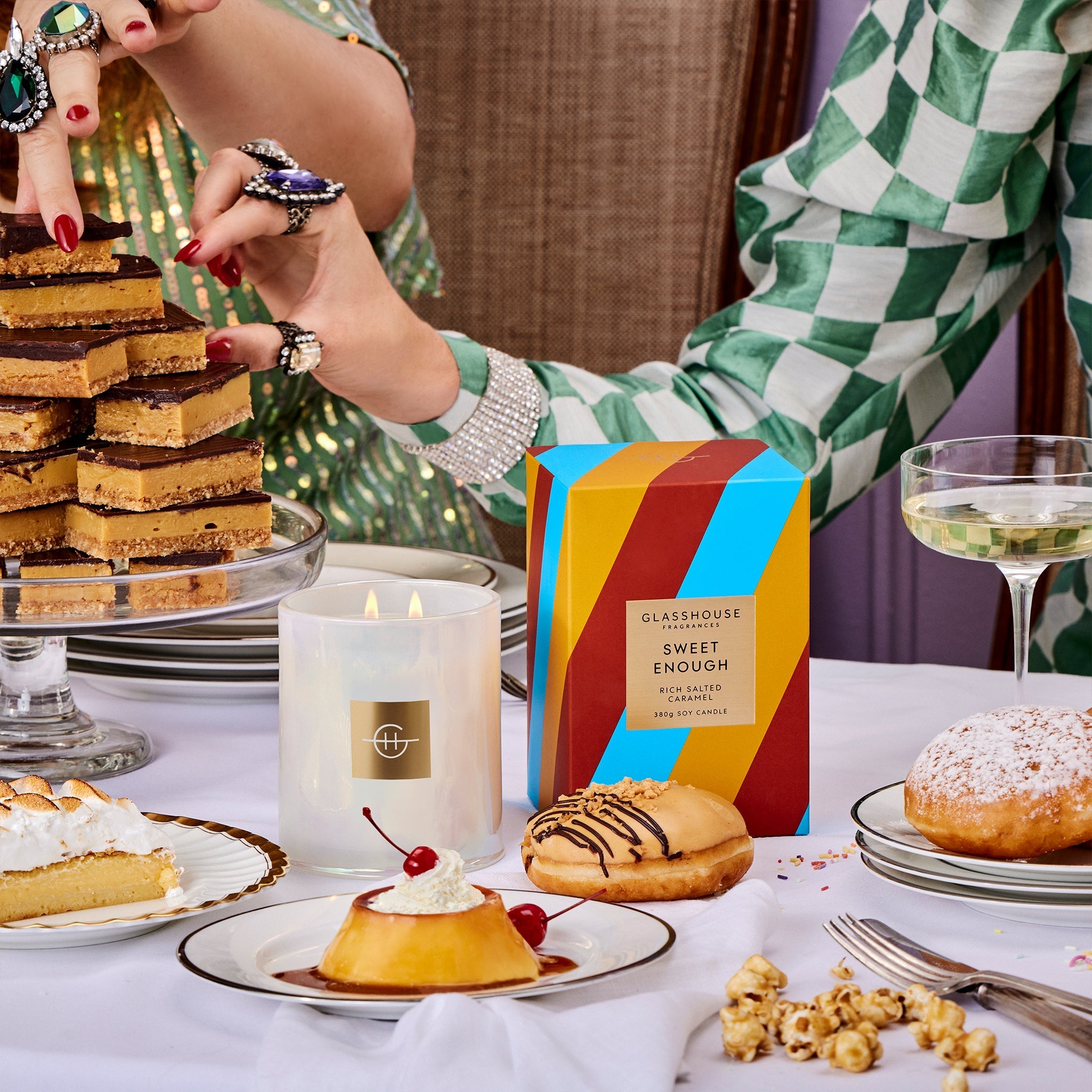 Glasshouse Fragrances Sweet Enough Salted Caramel 380g Soy Candle burning next to the box on a spread of caramel desserts